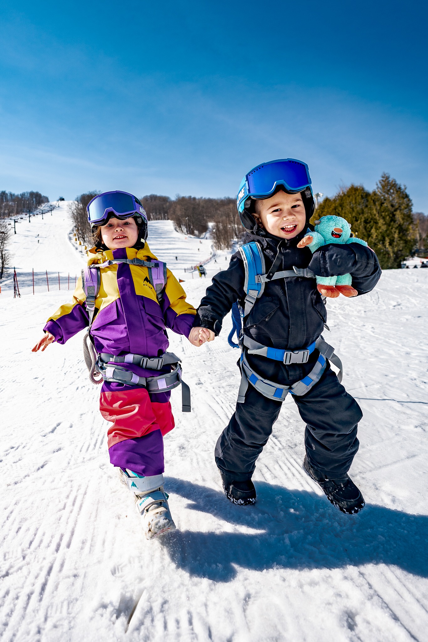 Two children walking hand-in-hand on snow wearing blue and lavender MDXONE harnesses, with a Yeti plush